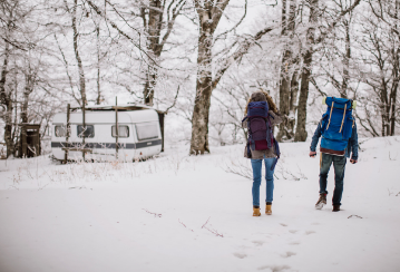 Two people carrying large hiking backpacks walk through the snowy woods towards their RV
