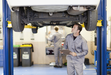 A mechanic stands underneath a car that's on a lift to inspect the undercarriage.