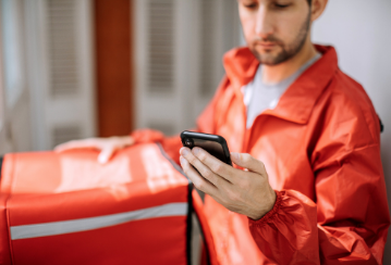 Delivery person stands with insulated food bag, looking at their phone on their way to deliver the food,