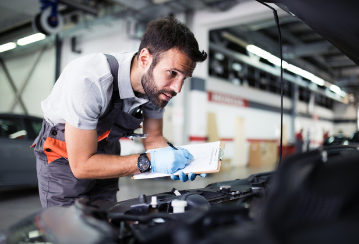 A mechanic looks under the hood of a car in a shop while taking notes on a clipboard.