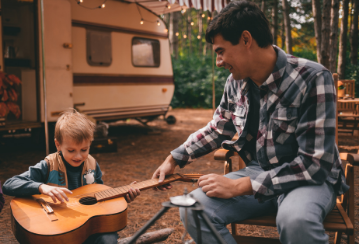 A father teaches his son to play guitar while sitting outside their camping trailer at a campsite.
