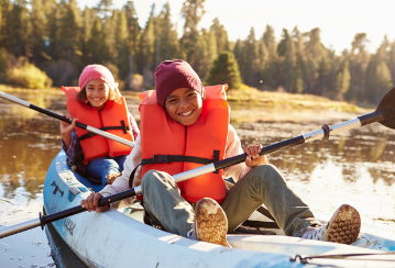 Two kids smile at the camera as they paddle a kayak in a lake.