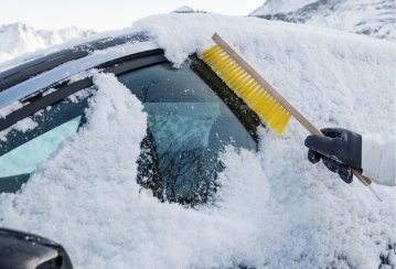 A gloved hand uses a snow brush to clear off a vehicle window.