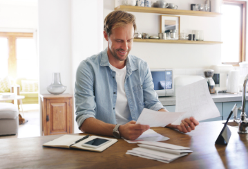 Man sitting at kitchen table looking at papers, with more papers and a notebook on the table.